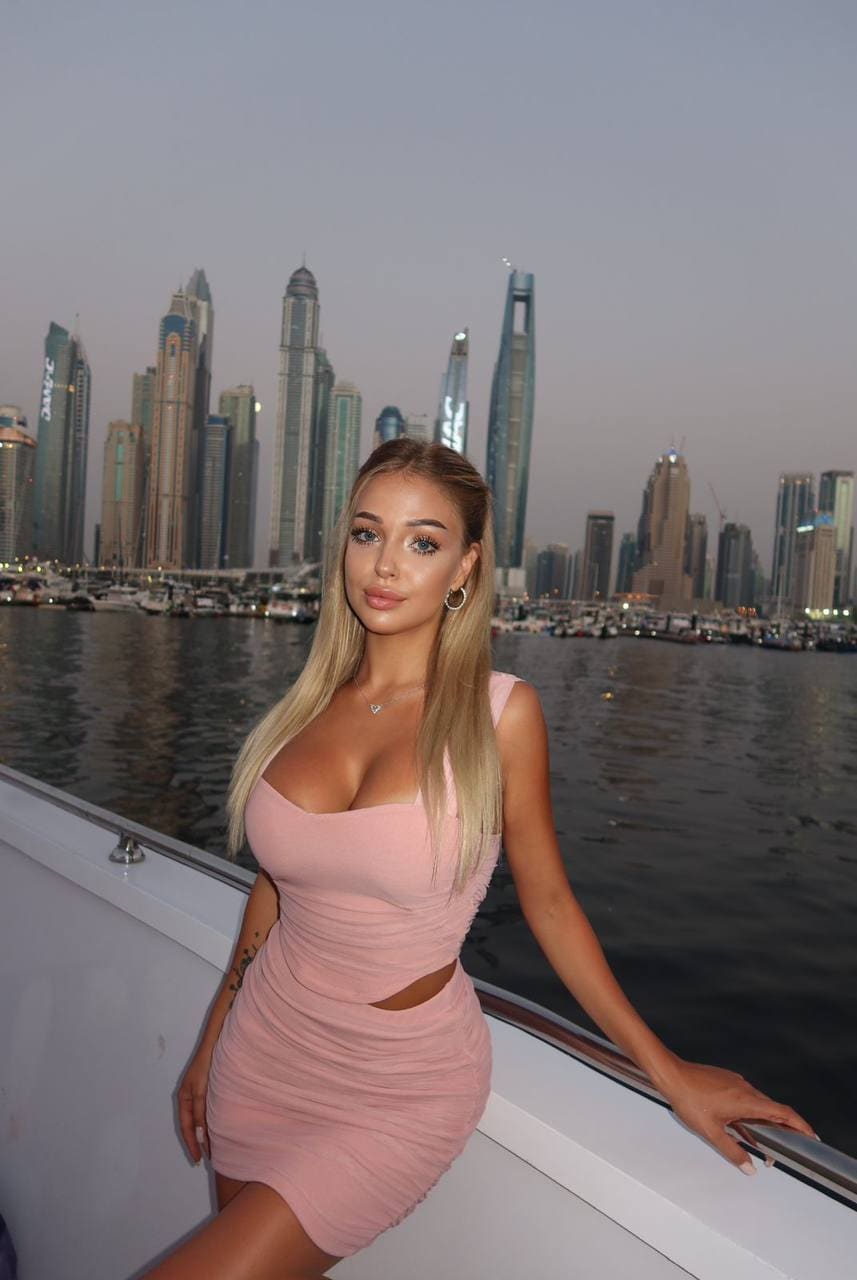 Blonde woman stands on a boat in a short pink dress with deep neckline and side cutouts, against a dusk city skyline and water.