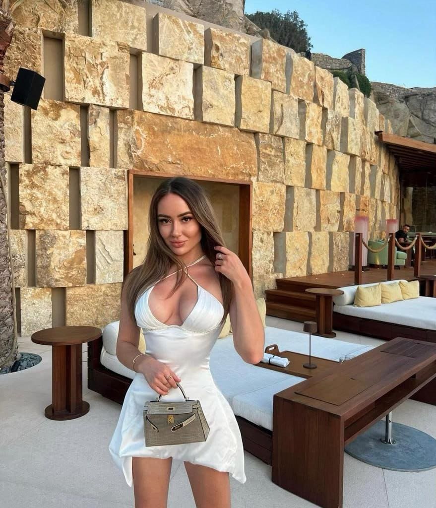 A woman in a short white halter-neck dress holds a structured handbag while standing before a stone wall with lounge seating in the background.