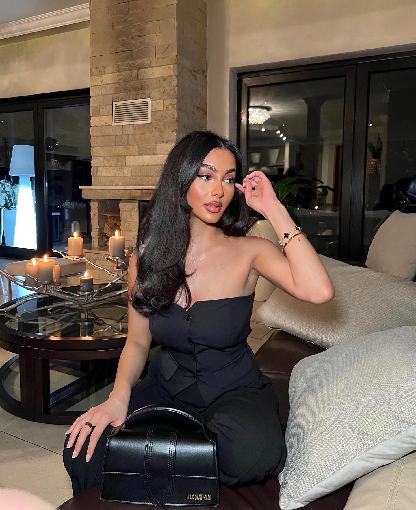 Aida sits on a living room couch in a black strapless top, holding a black handbag on her lap and touching her face.