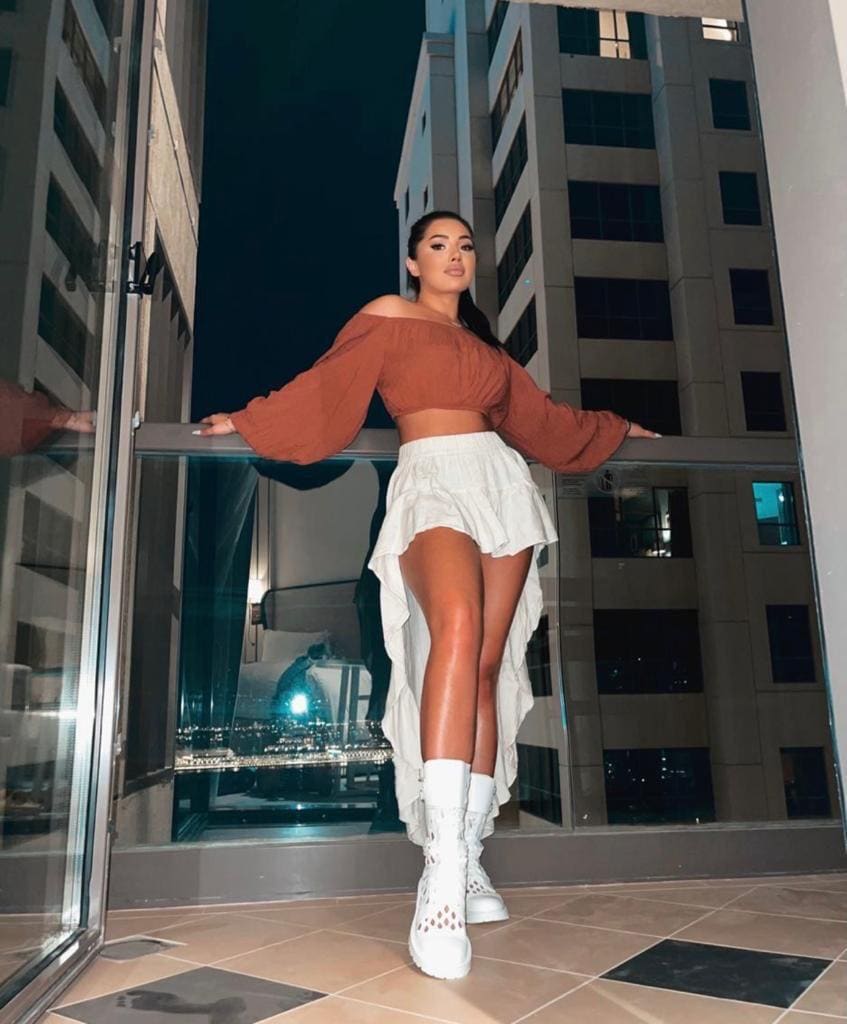 A woman stands on a tiled balcony at night in a rust crop top, white ruffled skirt, and lace-up boots, leaning on a glass railing near a building.