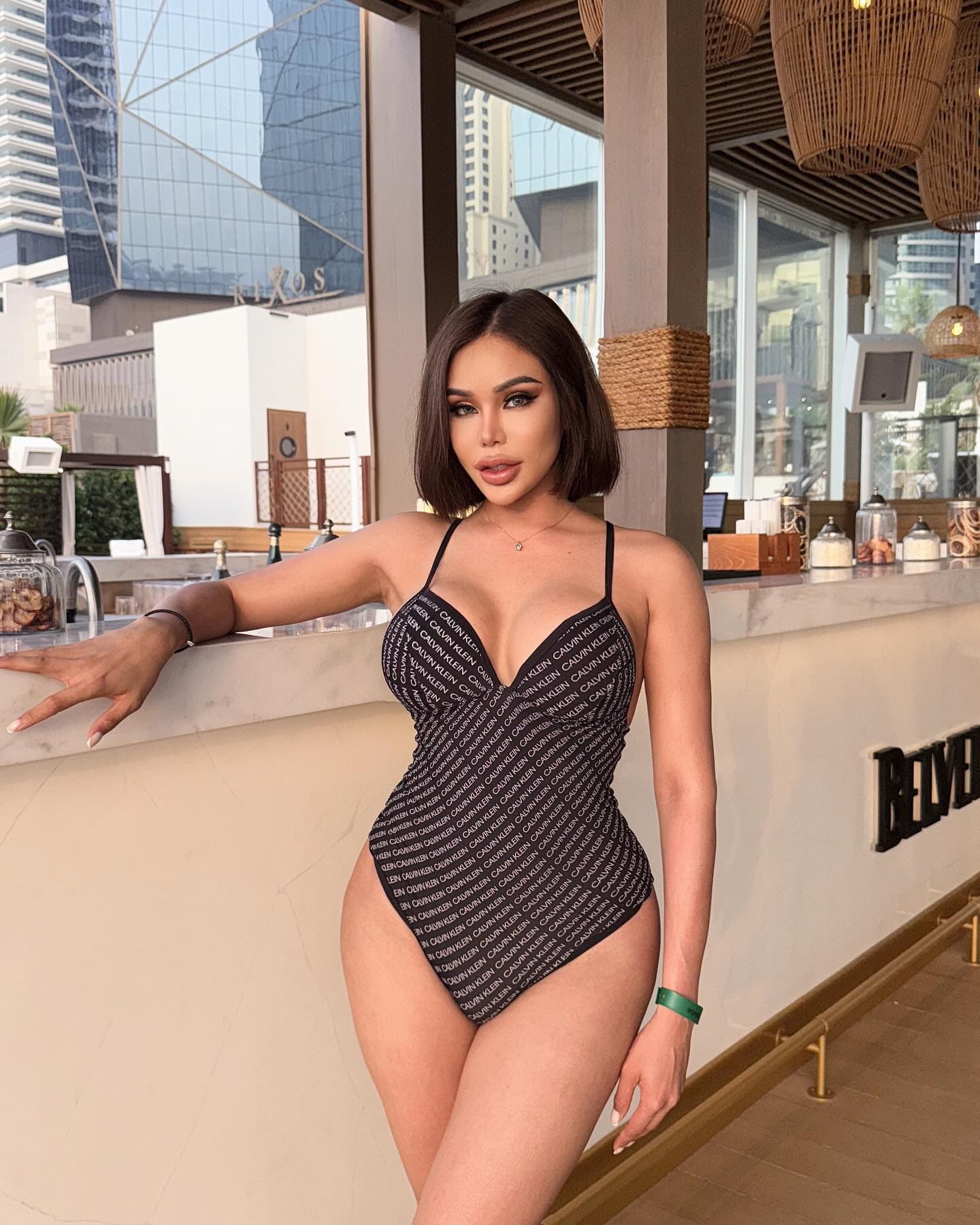 Nana stands at an indoor bar in a black swimsuit with white text, her arm on the marble counter with a city skyline behind.