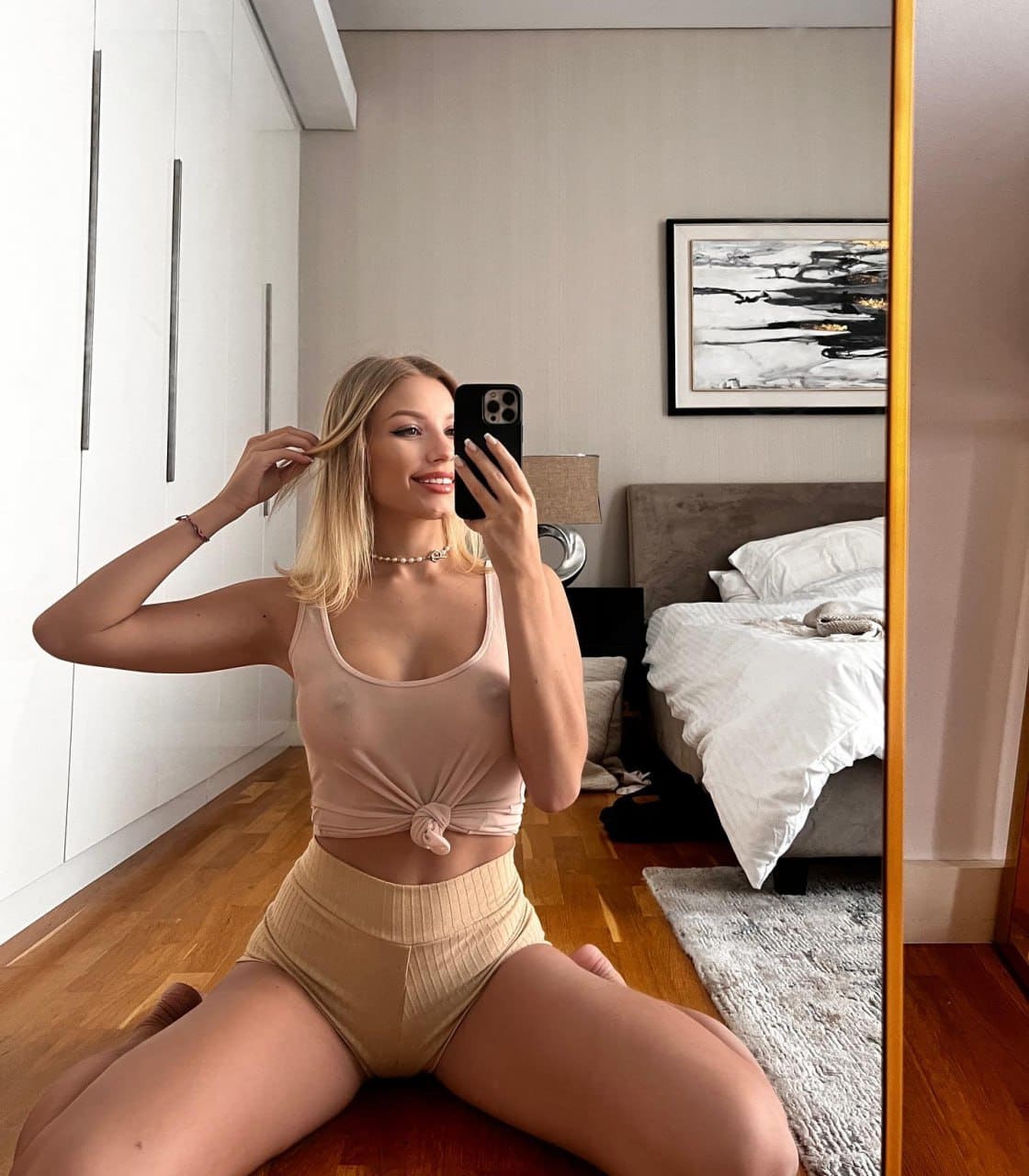 Sitting on a bedroom floor, a blonde woman in a pink knotted tank top and beige shorts takes a mirror selfie with a black smartphone.