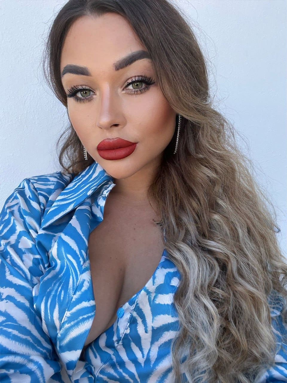 Anna with long wavy brown hair and red lipstick wears a blue and white patterned blouse with a deep neckline against a plain light wall.