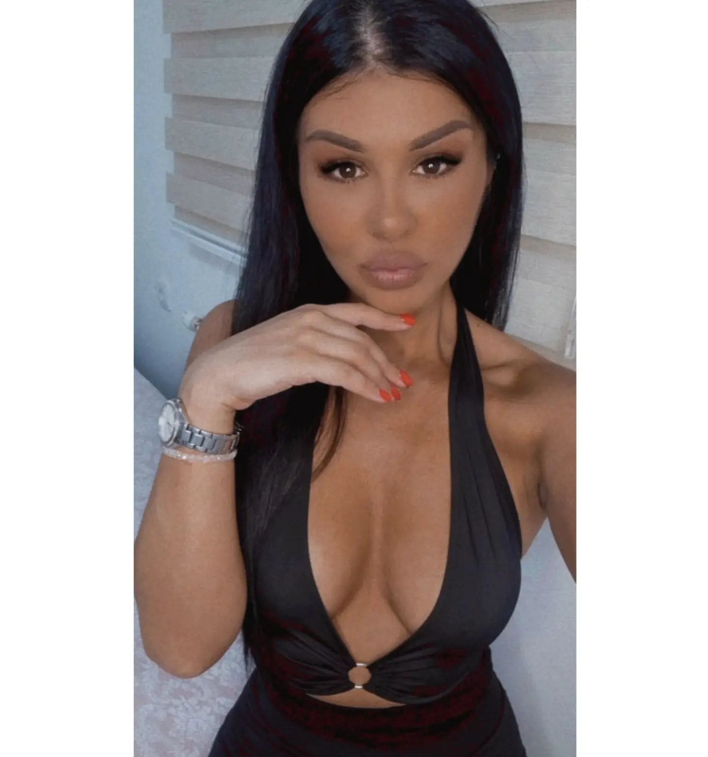 Ceharlott in a black halter-neck top with a deep neckline, hand near chin and silver watch visible, standing before white horizontal blinds.