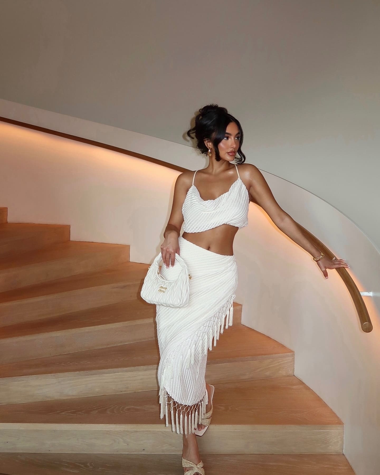 A woman with an updo stands on a wooden staircase in a white cropped top and fringed midi skirt, holding a white handbag.