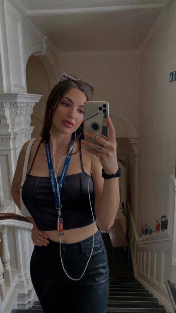 A woman takes a hallway mirror selfie wearing a black cropped tank, dark jeans and sunglasses on her head, holding a phone.