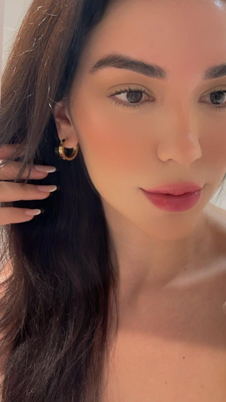 Close-up portrait of a woman with bare shoulders and a gold hoop earring, her hand raised near her ear against a light background.