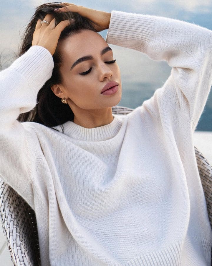 A woman sits in a woven chair outdoors near water, wearing a loose white knit sweater and gold earrings, with hands up to her head.