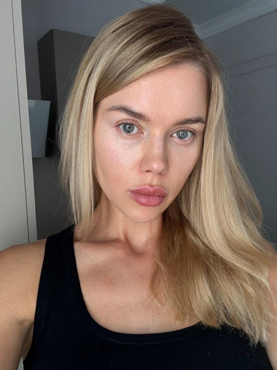 Stella with long blonde hair wears a black sleeveless top, standing indoors against a plain wall with a dark cabinet behind her.