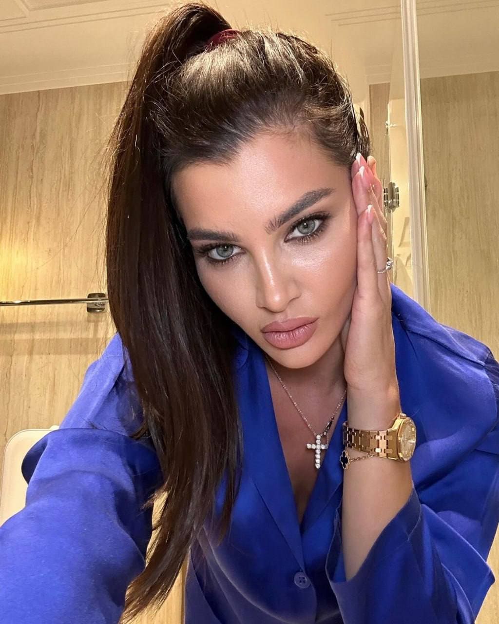 Juliette in a blue satin shirt with open buttons, hand on cheek, in a wood-paneled room.