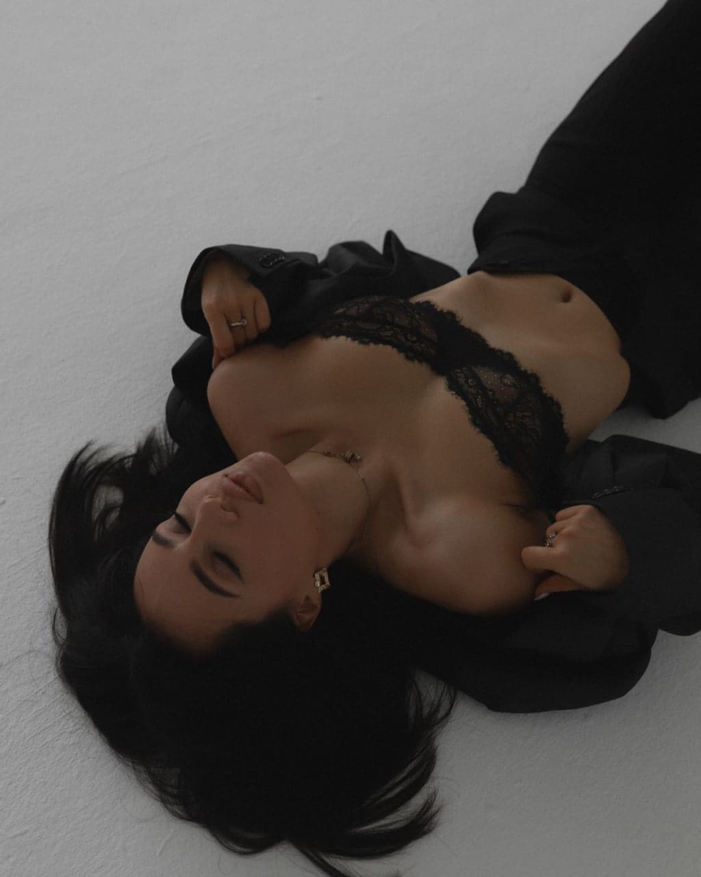 A woman lies on her back on a white surface, wearing a black lace bra and open jacket, with eyes closed and one hand near her chest.