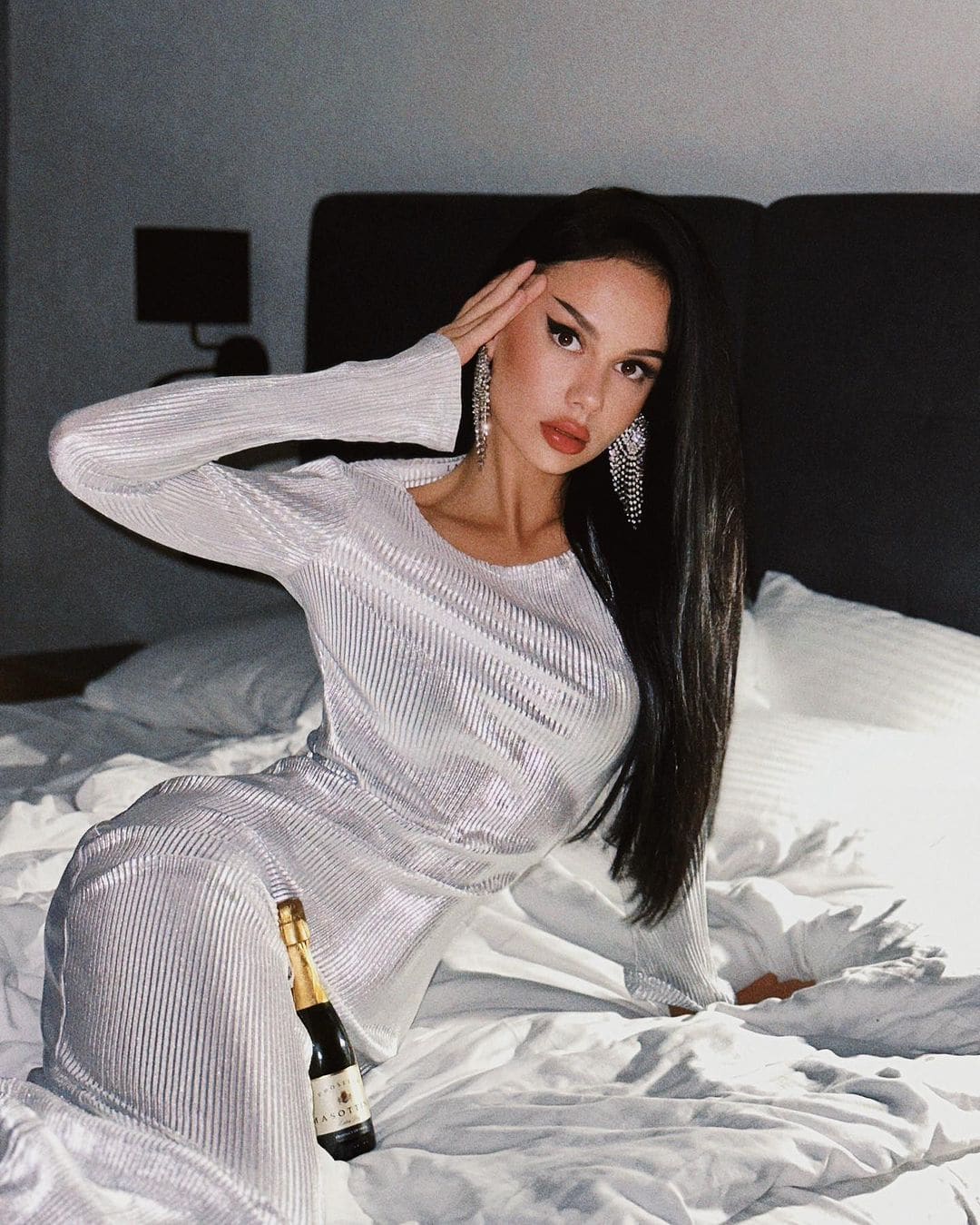 A woman in a ribbed light-colored dress and dangling earrings sits on a white bed, hand raised to her head, with a sparkling wine bottle between her legs.