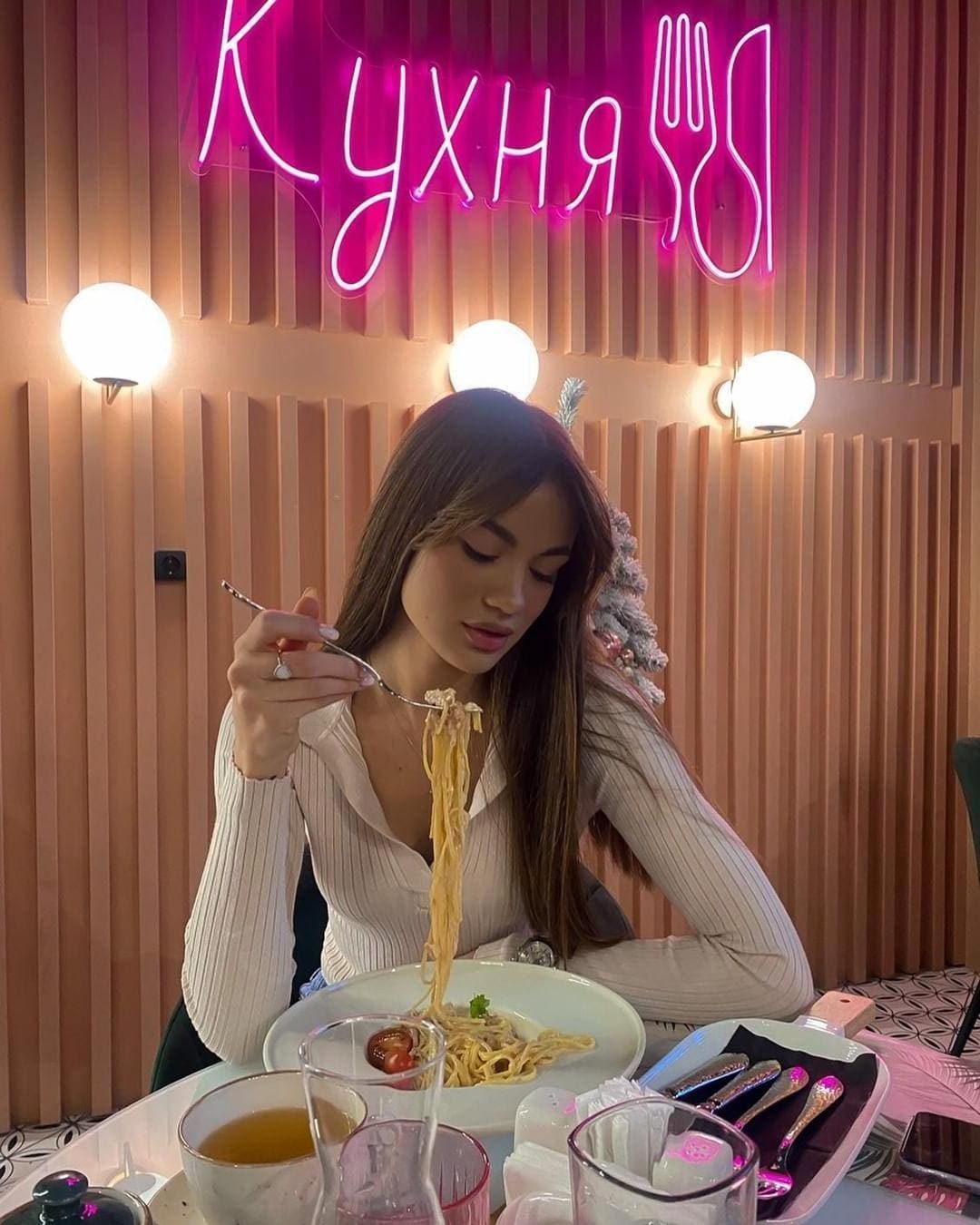 A woman in a white ribbed V-neck top sits at a restaurant table, lifting spaghetti to her mouth near a glowing neon sign on a slatted wall.