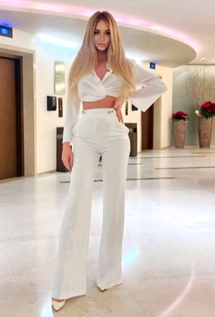 A woman stands in a bright lobby with marble floors, wearing a white cropped shirt and white wide-leg trousers.