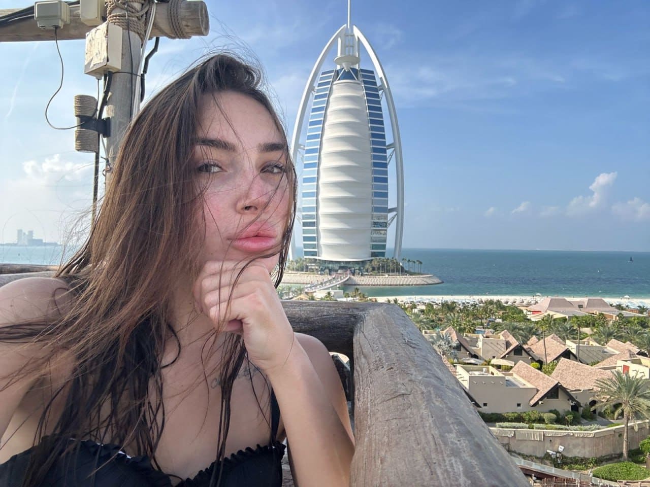 A woman with long wet brown hair rests her chin on her hand in a black strap top, standing on a wooden balcony overlooking a coastal resort and a sail-shaped skyscraper under a blue sky.