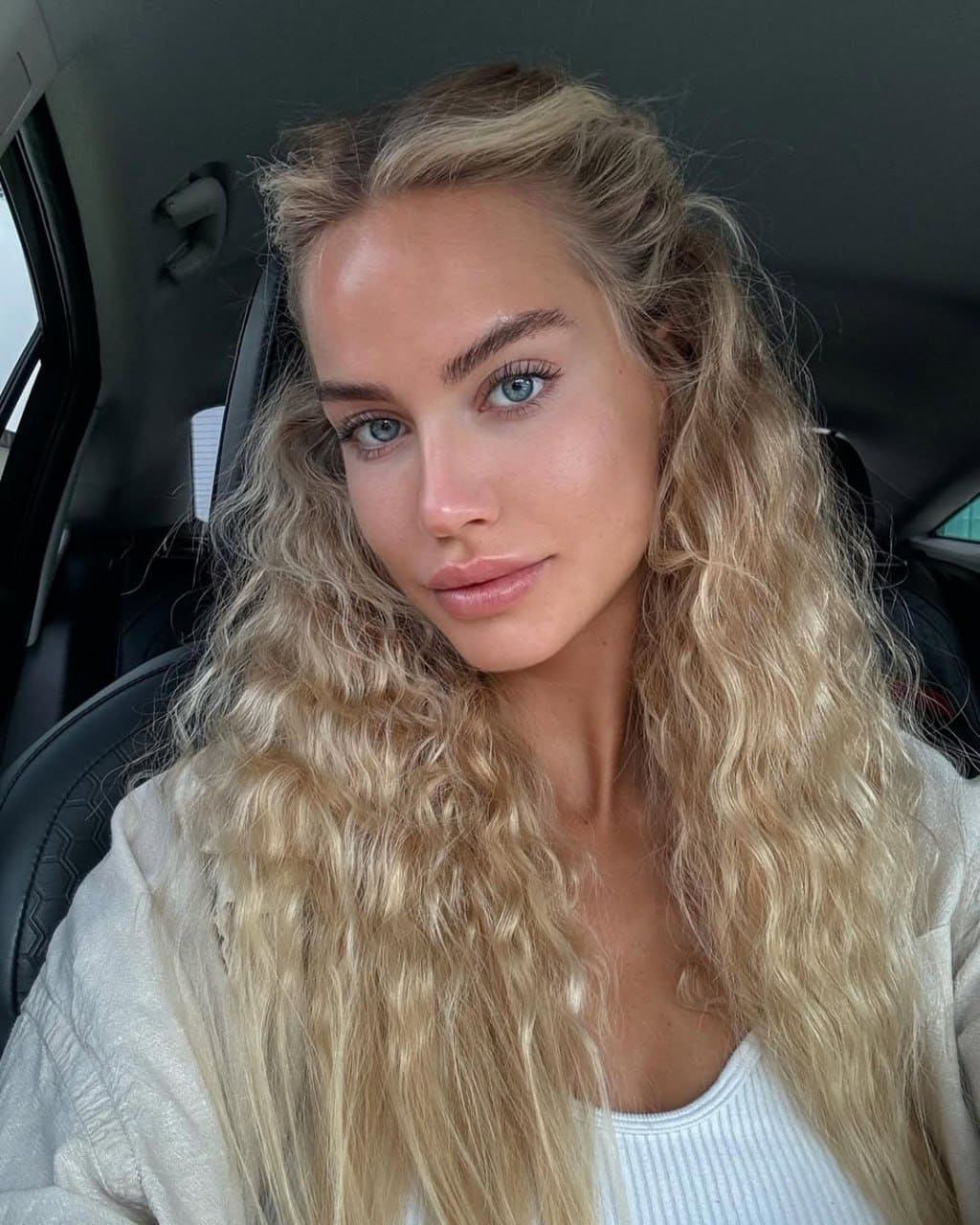 A woman with long wavy blonde hair sits in a car's driver seat, wearing a white ribbed tank top under a light cardigan, looking directly at the camera.