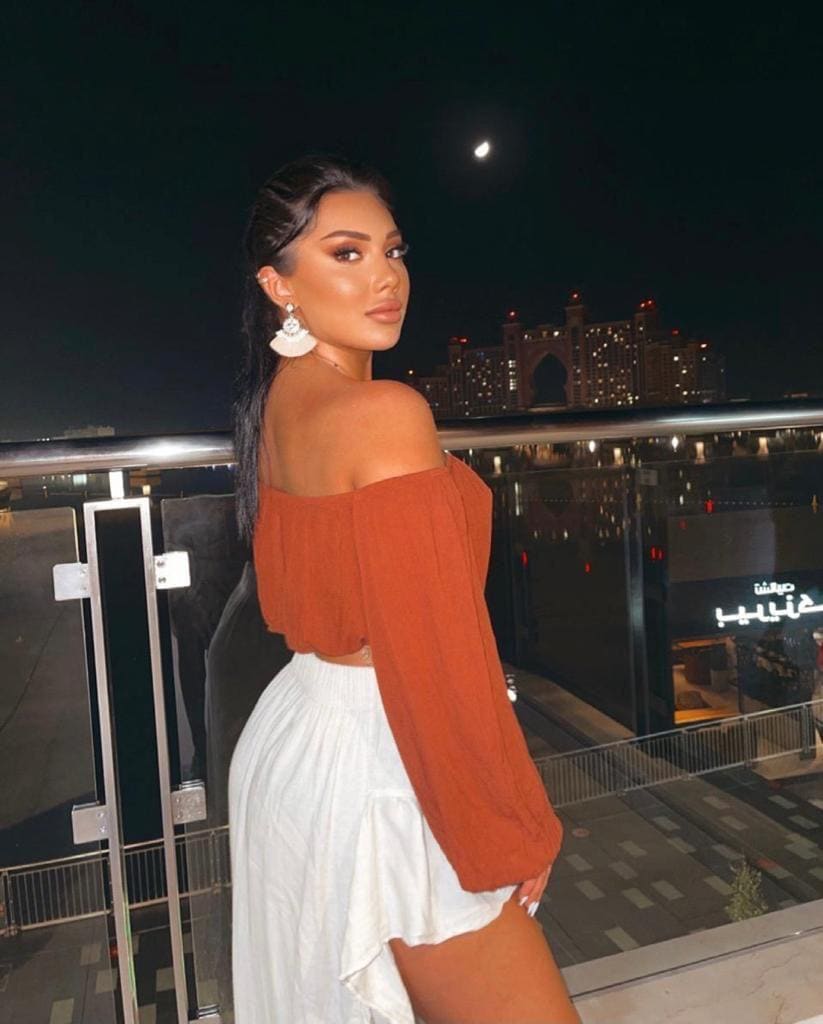 A woman stands on a rooftop at night in a rust off-the-shoulder crop top and white skirt, with a city skyline and crescent moon visible.