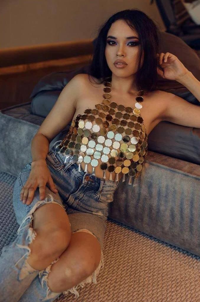 A woman sits on a gray couch wearing a metallic disc halter top and distressed denim shorts, one hand on her thigh and the other near her head.