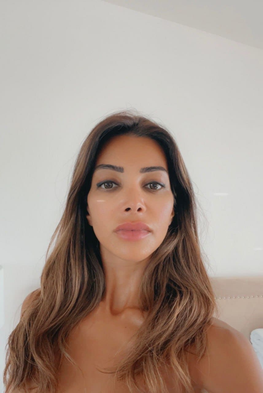Sara with long wavy brown hair, bare shoulders, in a close-up indoor portrait against a white wall and beige headboard.