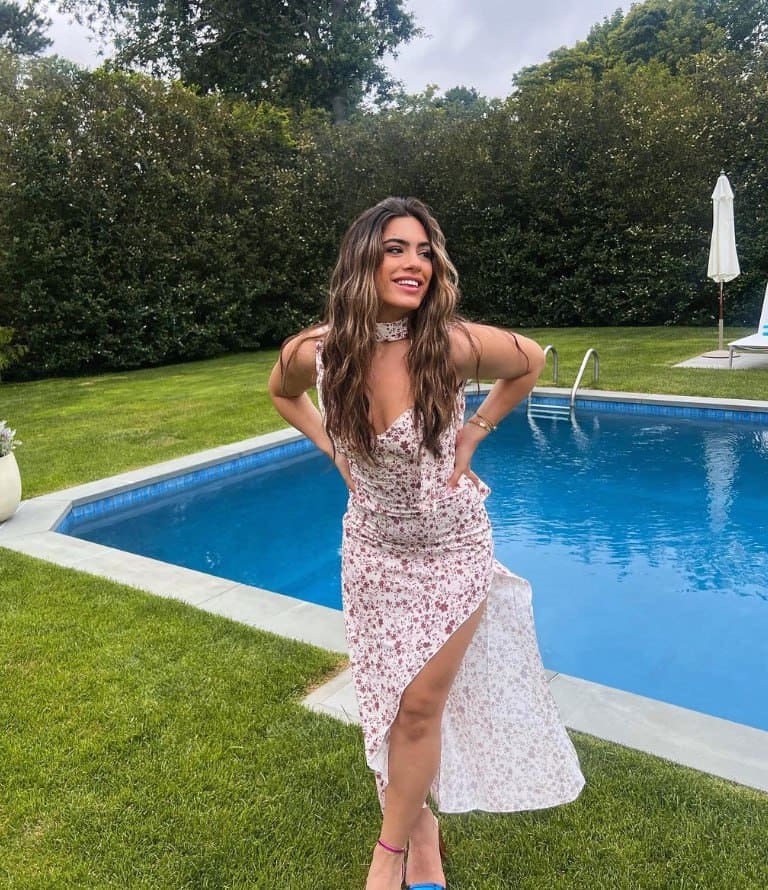 Caroline stands on a lawn by a pool in a floral dress with a high slit, hands on hips.