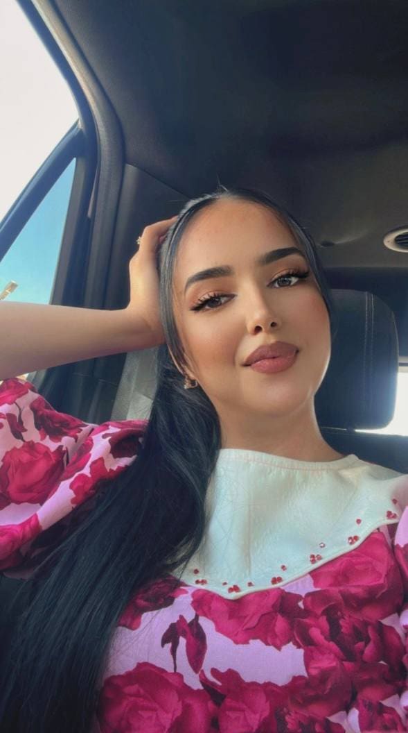 Astrid sits in a car passenger seat wearing a pink and red floral blouse with a white collar, her right hand raised to her head.