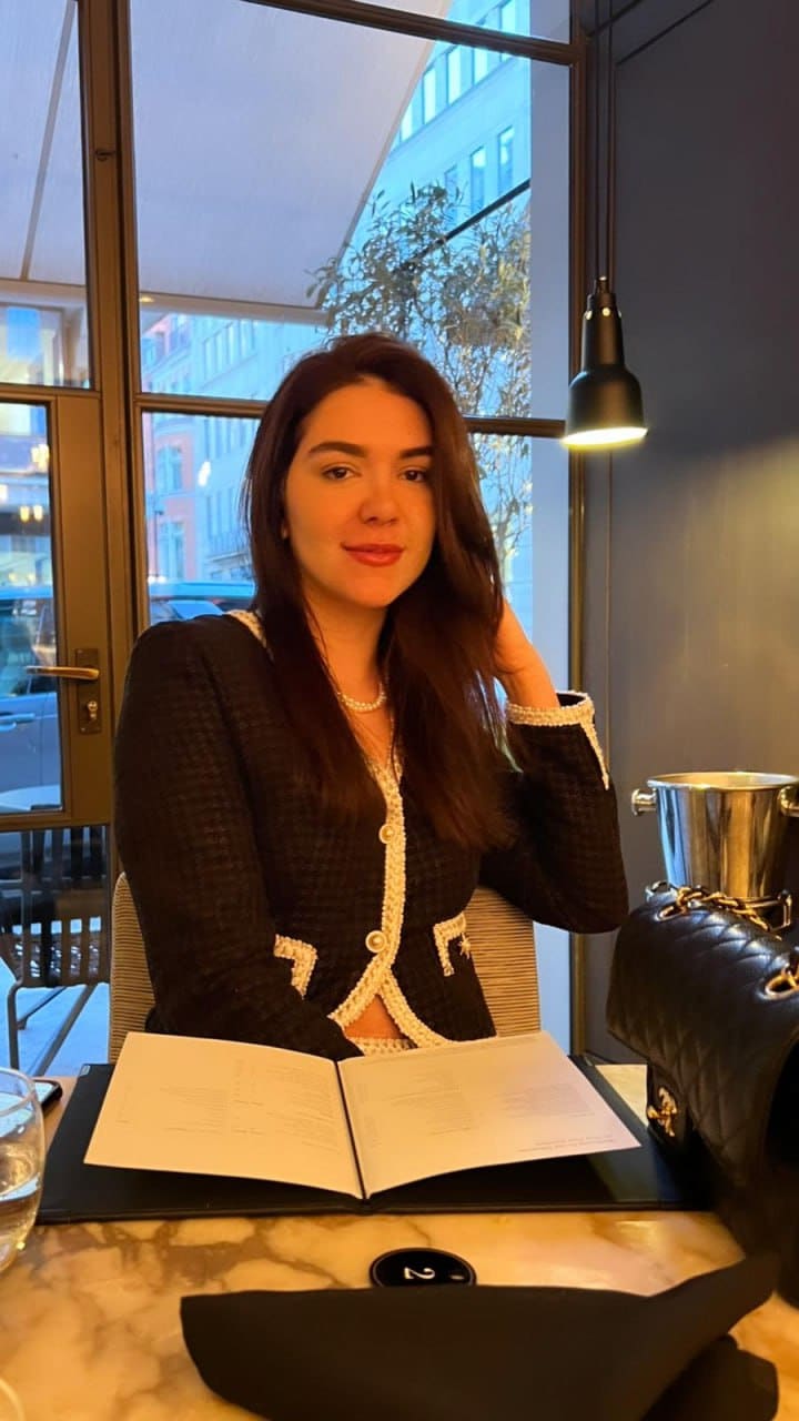 Beatriz sits at a marble restaurant table in a dark tweed jacket and pearl necklace, with an open menu and street view behind her.