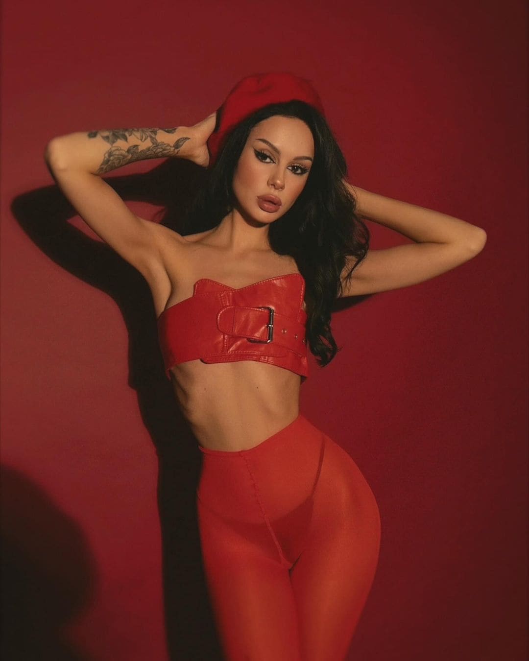 A woman in a red beret, red strapless bustier, and matching leggings poses with hands behind her head against a solid red studio backdrop.