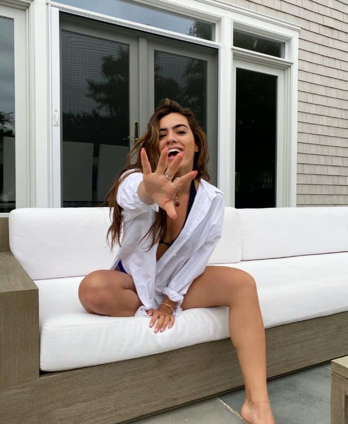 Sitting on a white sofa in an open white shirt over a dark bikini, she reaches a hand toward the camera.