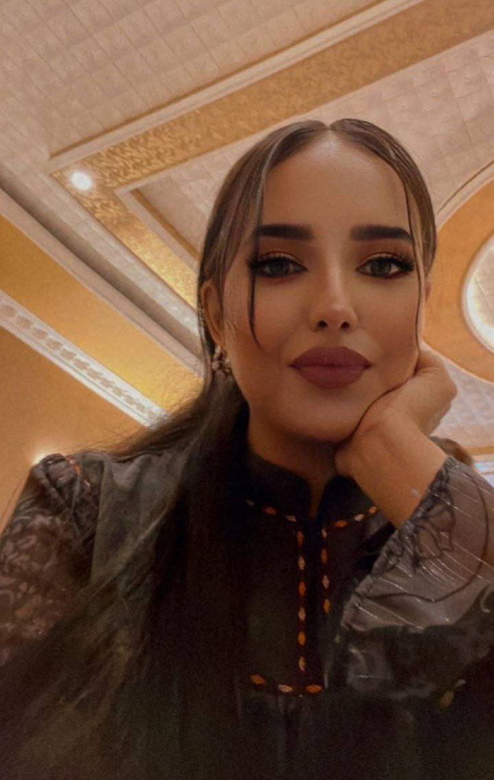 A woman with heavy eye makeup rests her chin on her hand, wearing a dark high-necked blouse with orange stitching against a patterned ceiling.