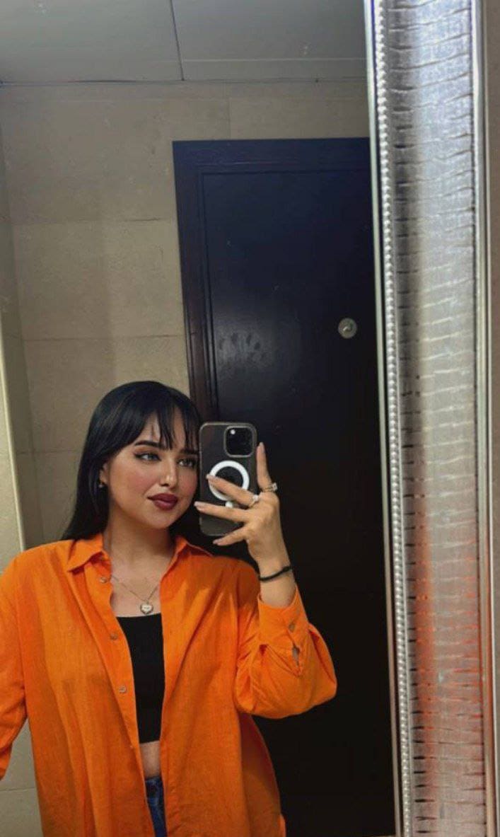 A woman with bangs takes a mirror selfie indoors, wearing an open orange button-up shirt over a black top and holding a smartphone.