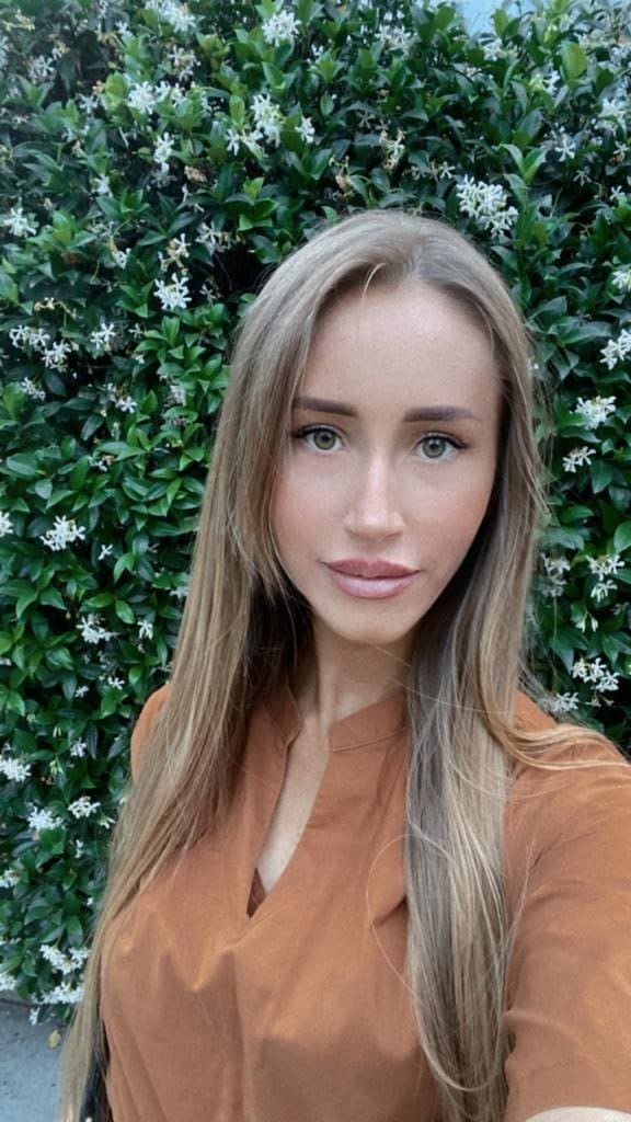 Nika stands before a green hedge with white flowers, wearing a rust-colored V-neck blouse in a close-up portrait.