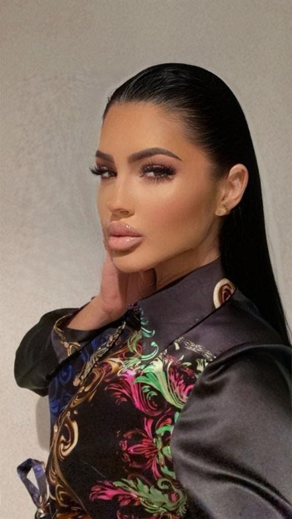 A woman with slicked-back hair poses in three-quarter view, wearing a high-collared floral blouse, her right hand resting near her chin.