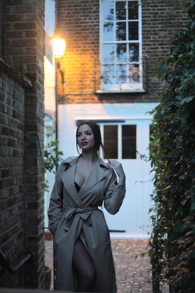 A woman stands in a brick alley at dusk wearing an open beige trench coat over a black bra and sheer tights, near a lit lamp.