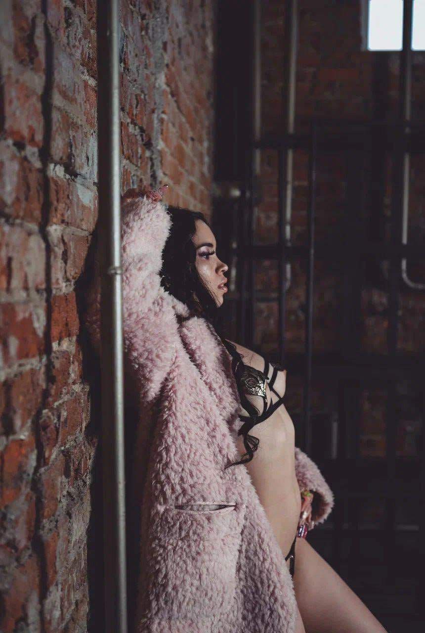 A woman stands in profile against a red brick wall, wearing an open pink fluffy coat over black strappy lingerie, with one arm raised along the wall.