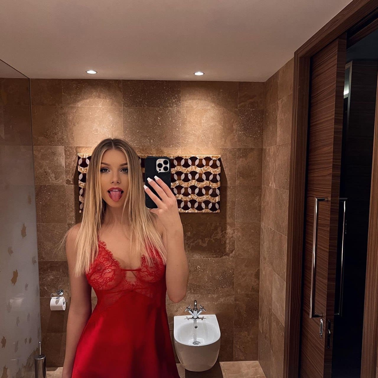 In a bathroom, a blonde woman in a red lace-trimmed slip dress sticks her tongue out while taking a mirror selfie, with patterned tiles visible.