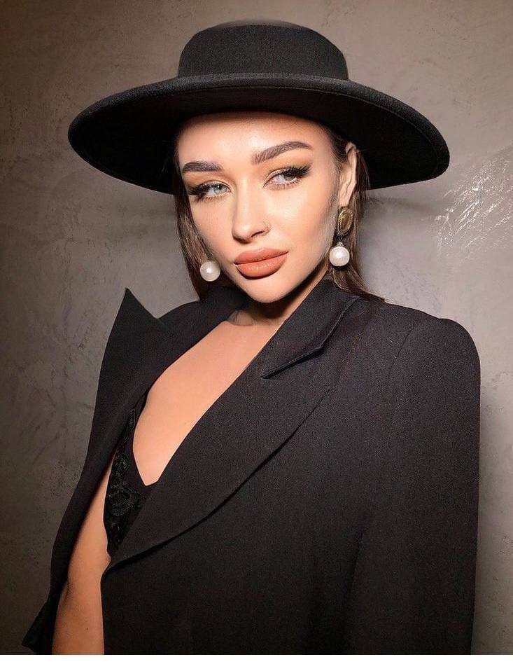 A woman in a wide-brimmed black hat and black blazer over a black lace top poses against a textured gray wall.