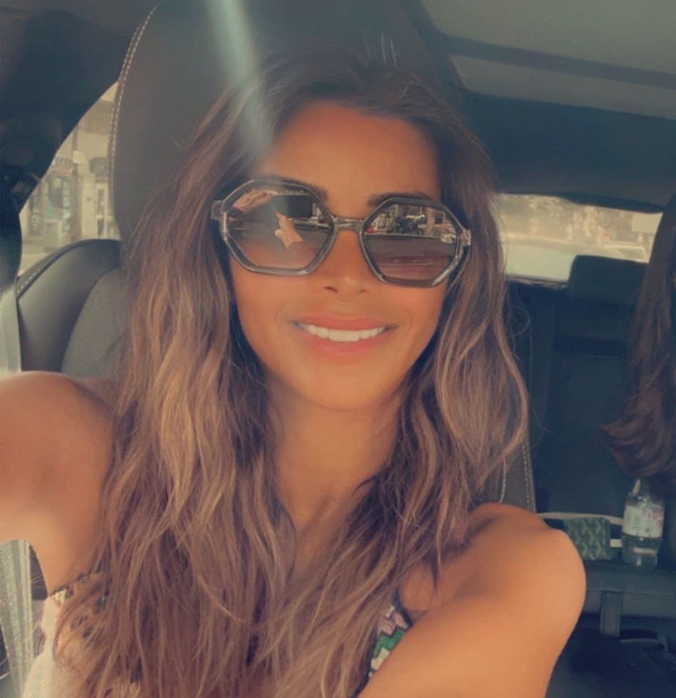 Woman with wavy brown hair, octagonal sunglasses, and floral top smiles while seated in a car back seat.