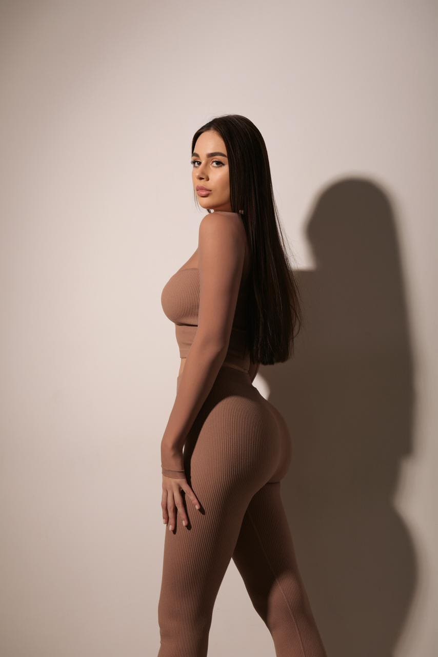 Profile view of a woman in a brown ribbed crop top and high-waisted leggings, hand on hip, against a plain light wall.