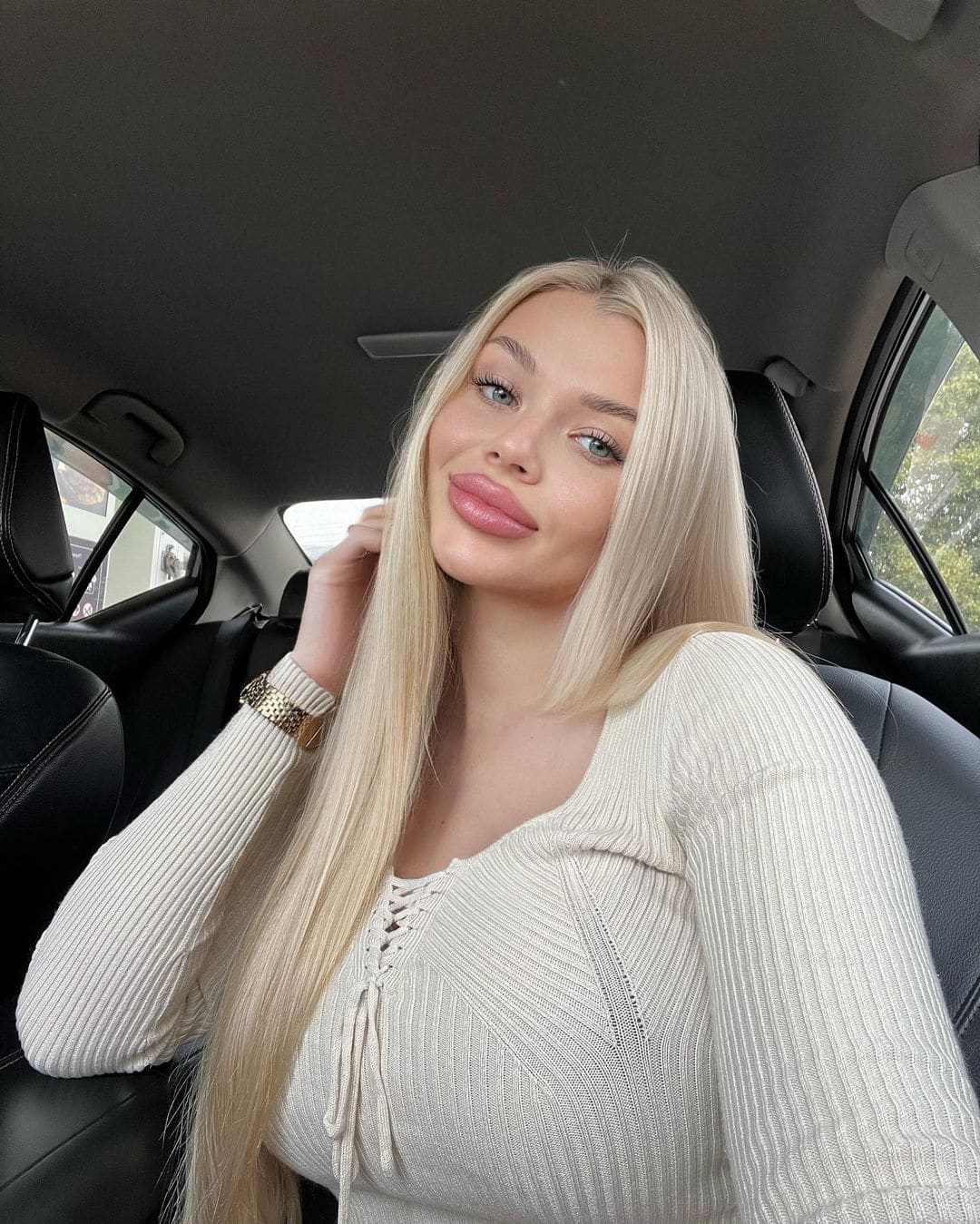 Assol sits in a car back seat, wearing a cream lace-up ribbed top and gold watch, with one hand near her neck.
