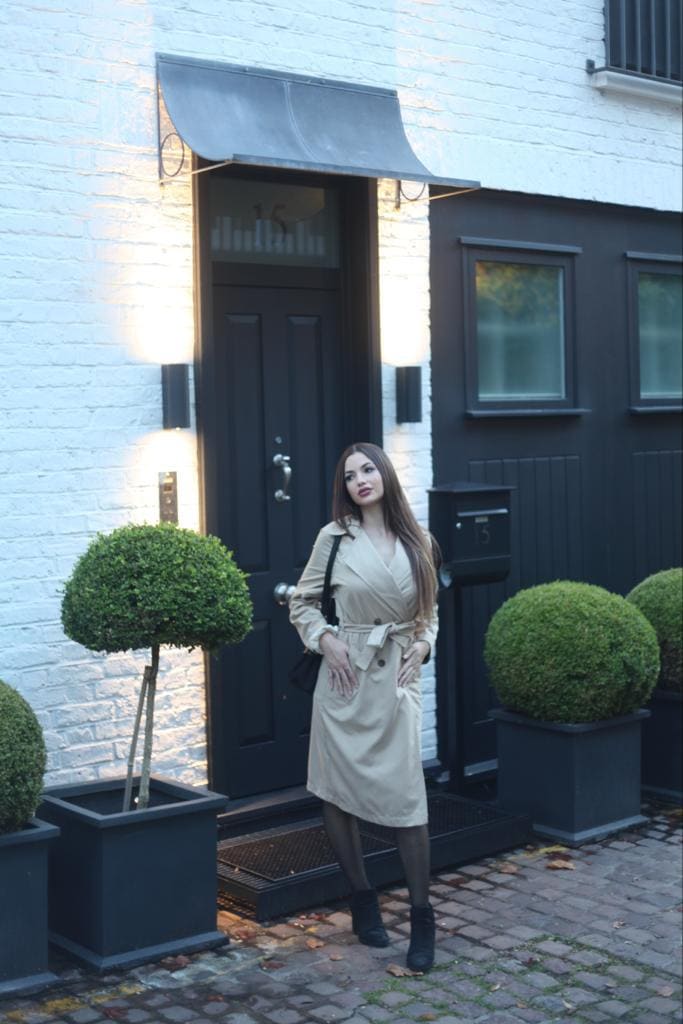A woman in a belted beige trench coat, dark tights and ankle boots stands on a brick walkway with hands on hips and a shoulder bag.