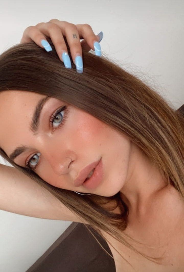 Close-up portrait of a woman with blue eyes and a dark strapless top, resting a hand with light blue nails on her head.