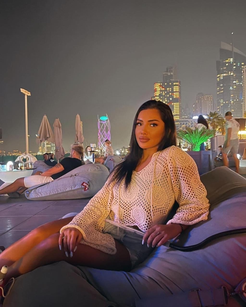 A woman sits on a gray beanbag at a rooftop venue at night, wearing a cream open-knit cardigan and denim shorts, with a city skyline behind.