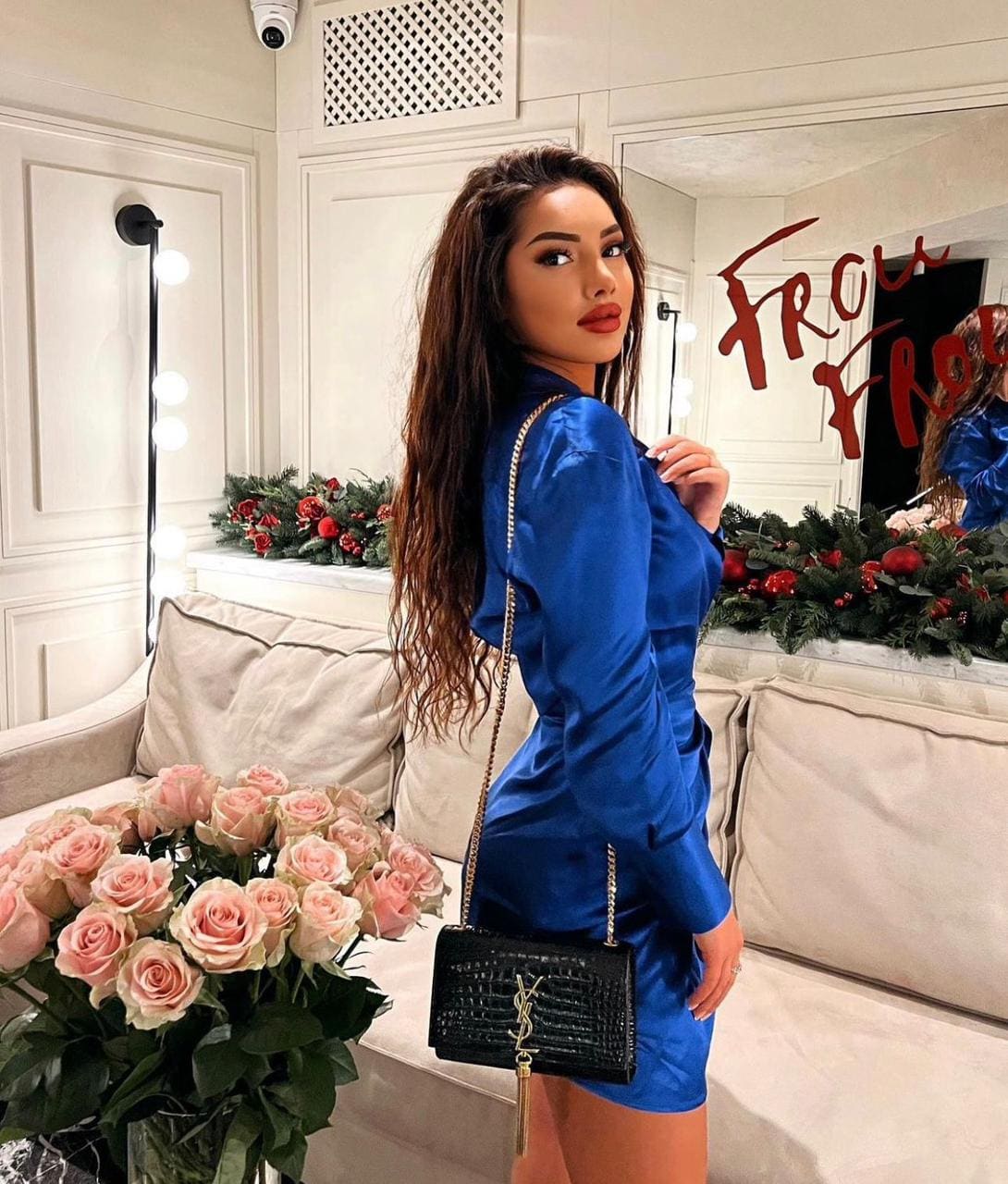 A woman stands in a room with white paneled walls, wearing a blue satin mini dress and holding a black quilted bag, beside a sofa and pink roses.