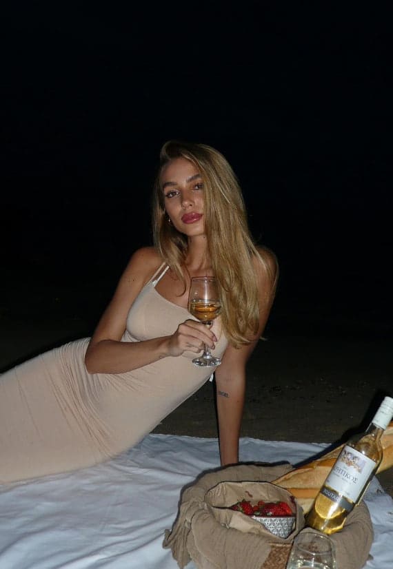 A woman in a beige dress lies on white sheets at night, holding a glass of white wine, with a bottle and strawberries nearby.