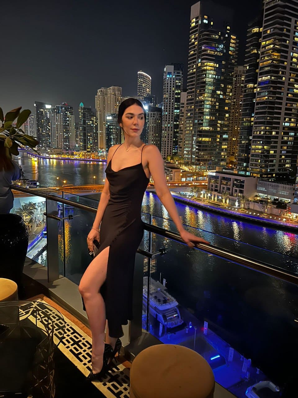 A woman in a black slip dress and heels leans on a rooftop glass railing at night, overlooking a city skyline and river.