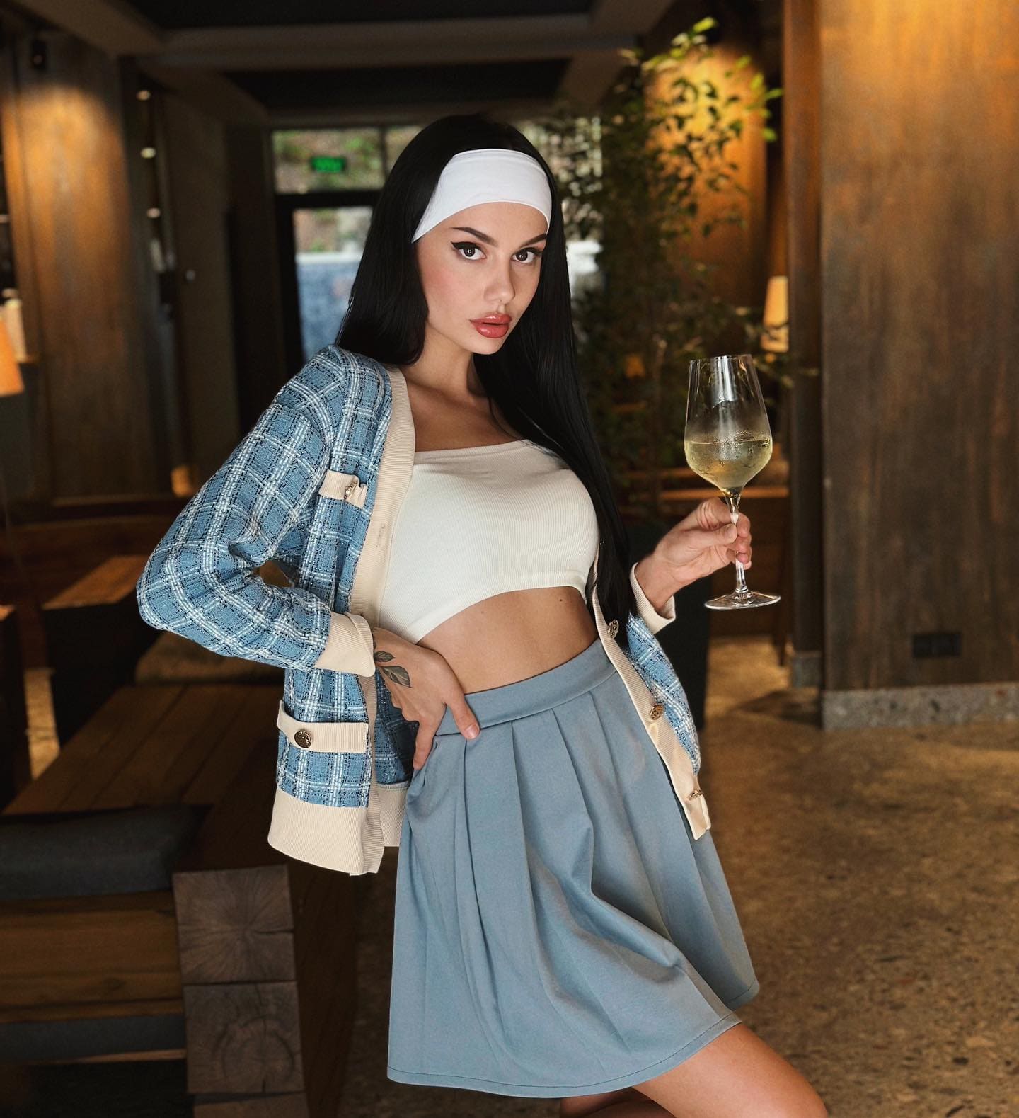 Lily stands indoors holding white wine, wearing a white crop top, blue plaid cardigan, and light blue pleated skirt with one hand on her hip.