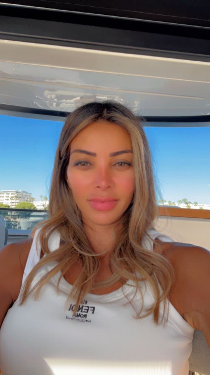 Blonde woman in white sleeveless top takes a selfie on a boat, with blue sky and distant buildings visible.