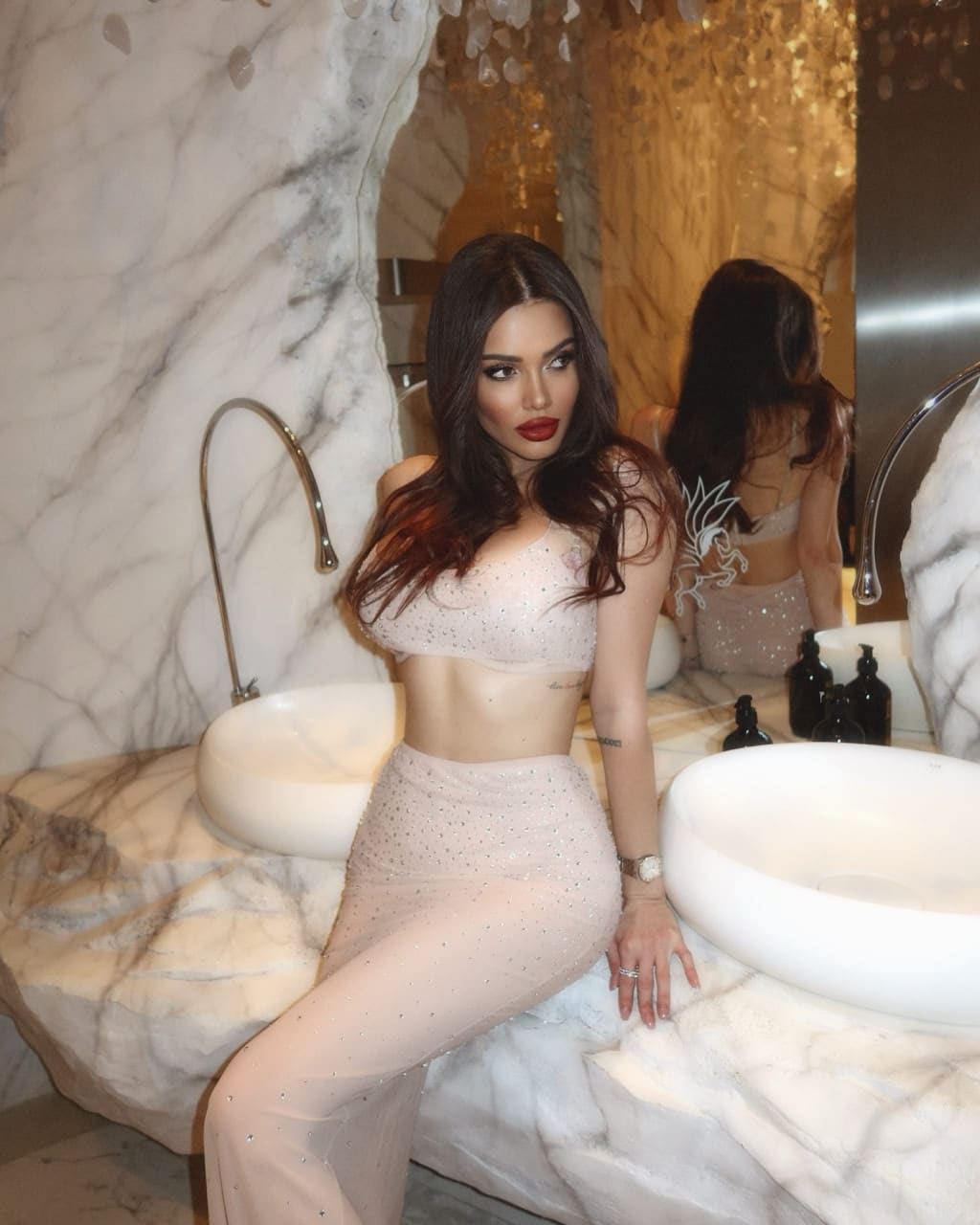 A woman in a sparkly two-piece outfit sits on a marble counter beside a white vessel sink and gold faucet in a tiled bathroom.