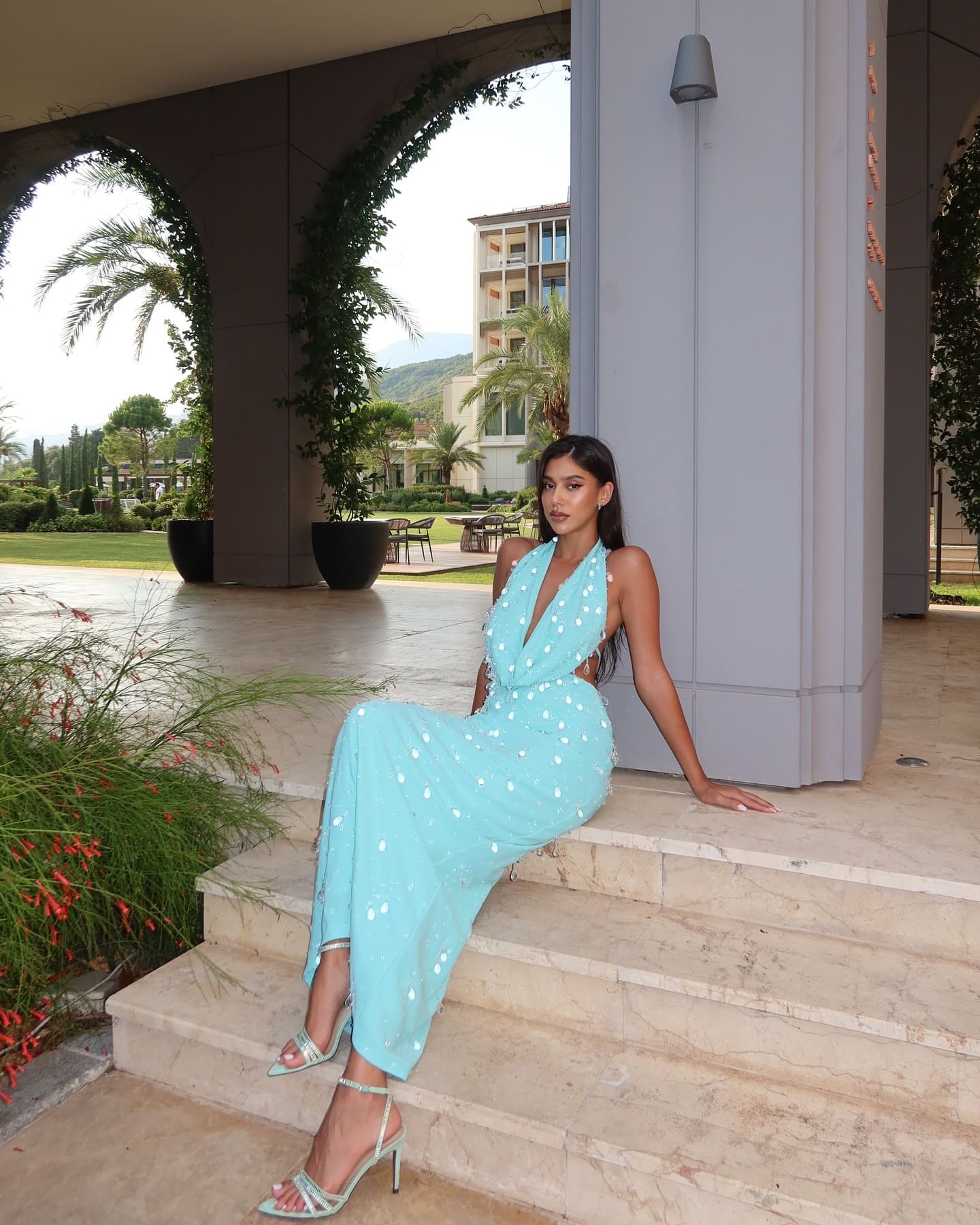 Seated on stone steps, a woman wears a light blue dotted halter gown with side cutouts and strappy heels in a resort setting.