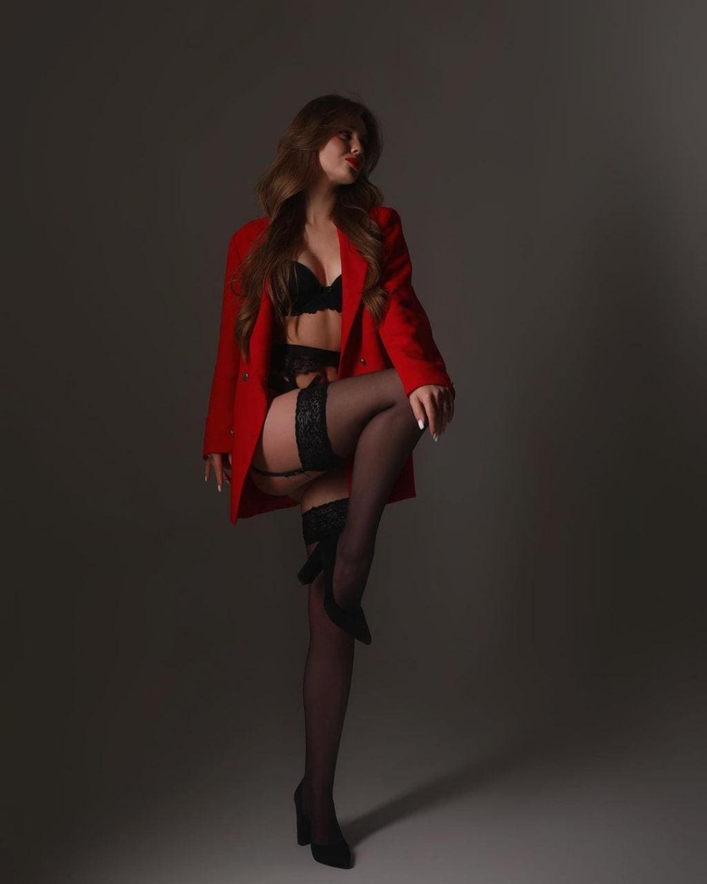 A woman poses in a studio wearing an open red blazer, black lingerie, and thigh-high stockings, with one leg raised.